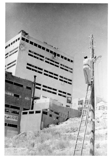 עובד חברת החשמל מתקן קו חשמל בחיפה בזמן המלחמה. על הבניין נראים סימני הפגיעה